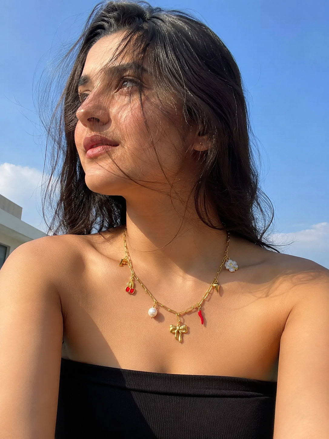 A woman outdoors wearing a delicate gold necklace with small charms including a white pearl, a red flower, and a gold cross pendant, paired with a black strapless top against a blue sky.