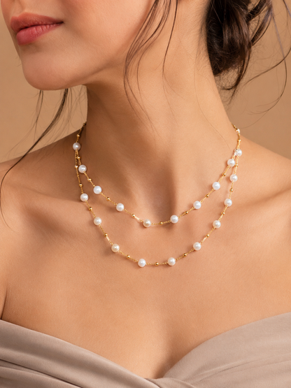 A close-up of a model wearing a double-strand gold necklace with small white pearls, styled with a taupe off-the-shoulder top against a warm beige background.