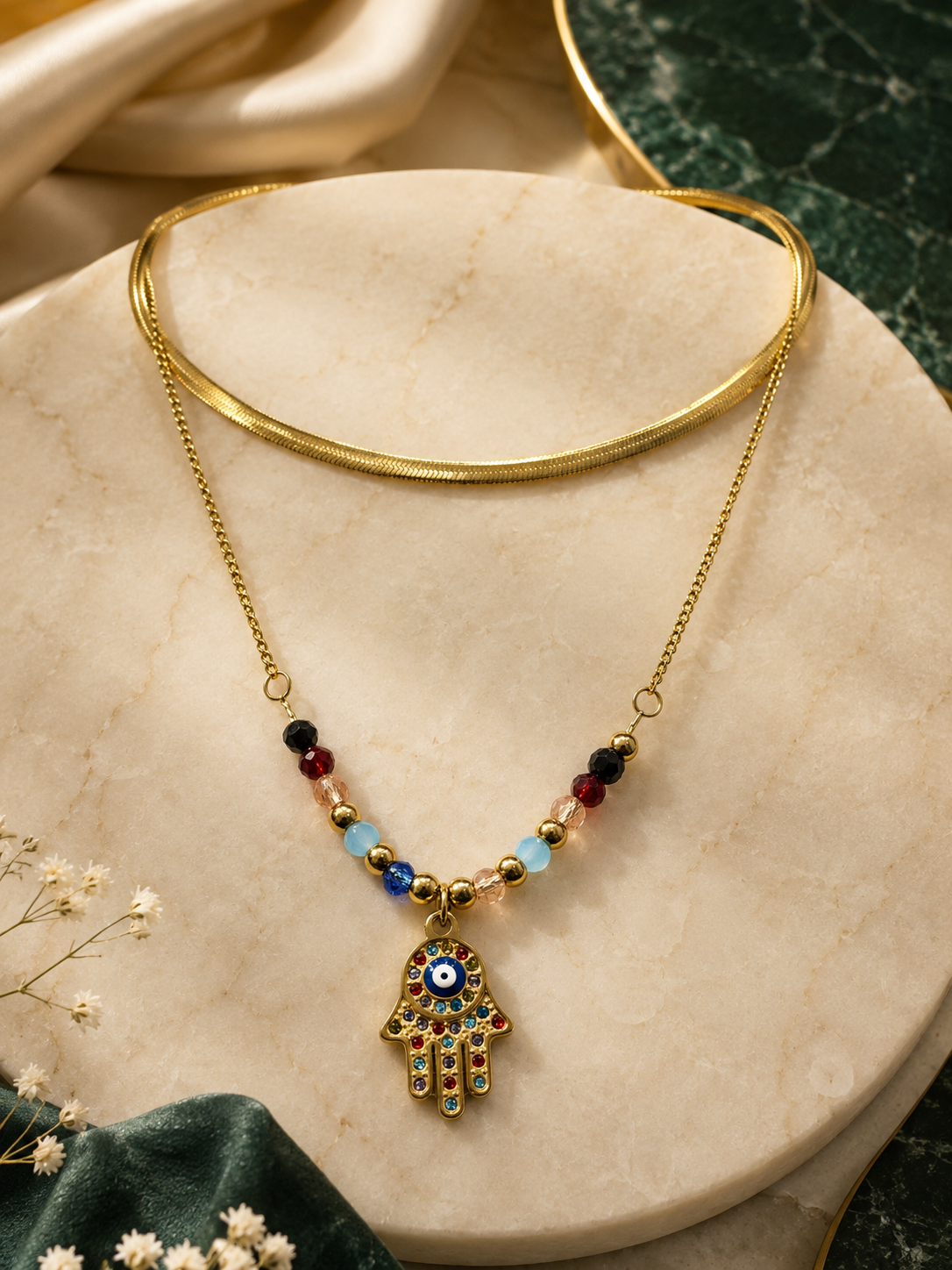 Gold-tone Hamsa hand pendant necklace with colorful evil eye details and small beads, displayed on a beige marble stand with a gold choker behind it.