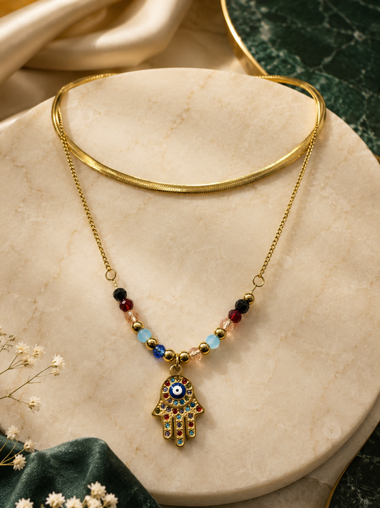 Gold-tone Hamsa hand pendant necklace with colorful evil eye details and small beads, displayed on a beige marble stand with a gold choker behind it.
