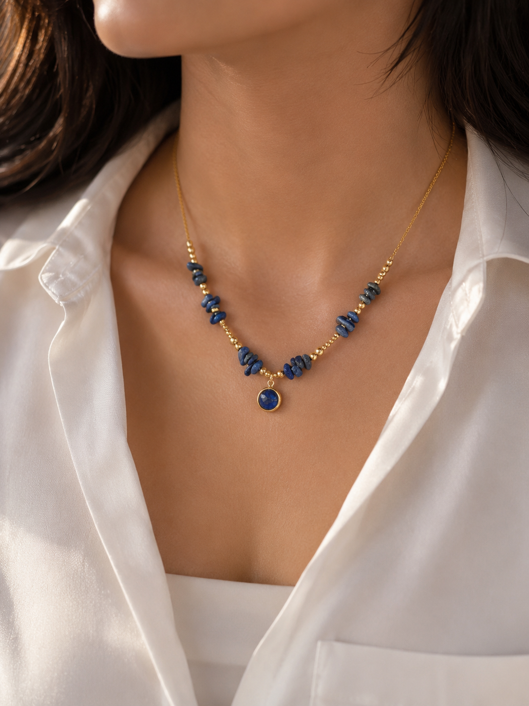 A close-up of a woman wearing a delicate gold necklace with blue stone beads and a round blue pendant, styled with a white collared shirt.