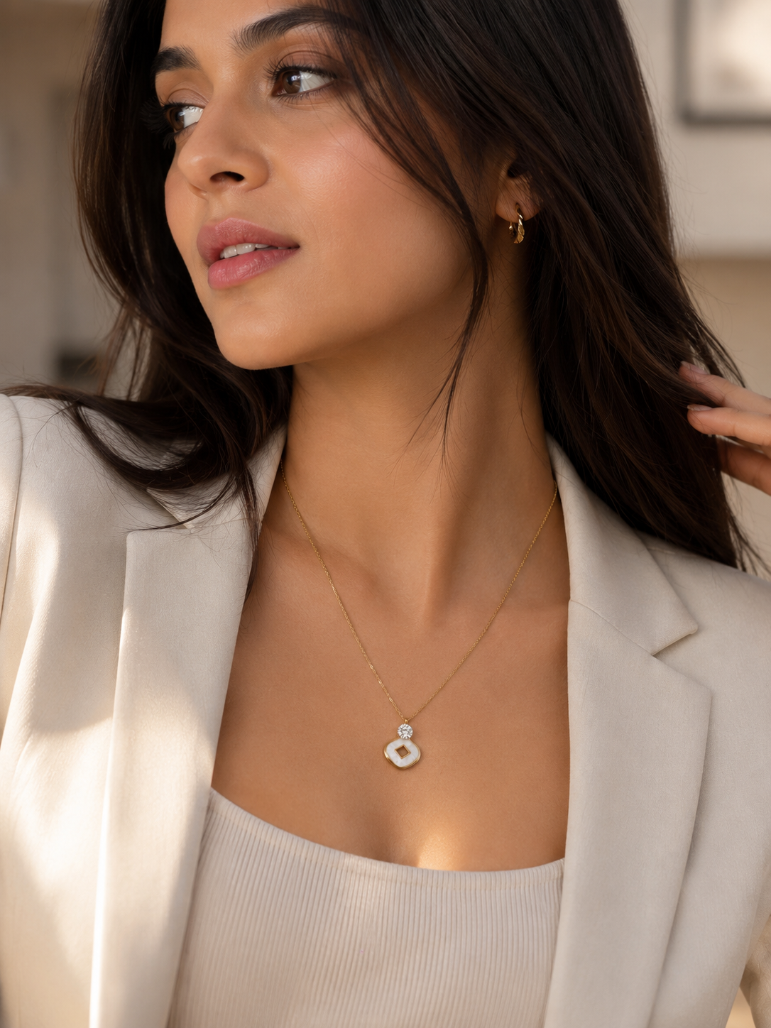 A woman wearing a delicate gold chain necklace with a white circular pendant, styled with a cream-colored blazer and top.