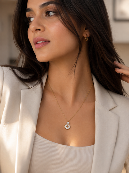 A woman wearing a delicate gold chain necklace with a white circular pendant, styled with a cream-colored blazer and top.