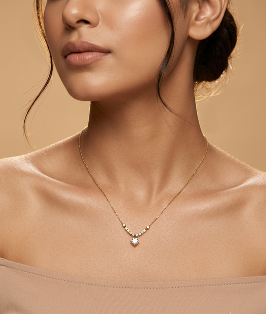 A close-up lifestyle shot of a model wearing a delicate gold necklace with a small heart-shaped crystal pendant and a row of tiny clear stones, set against a warm beige background.